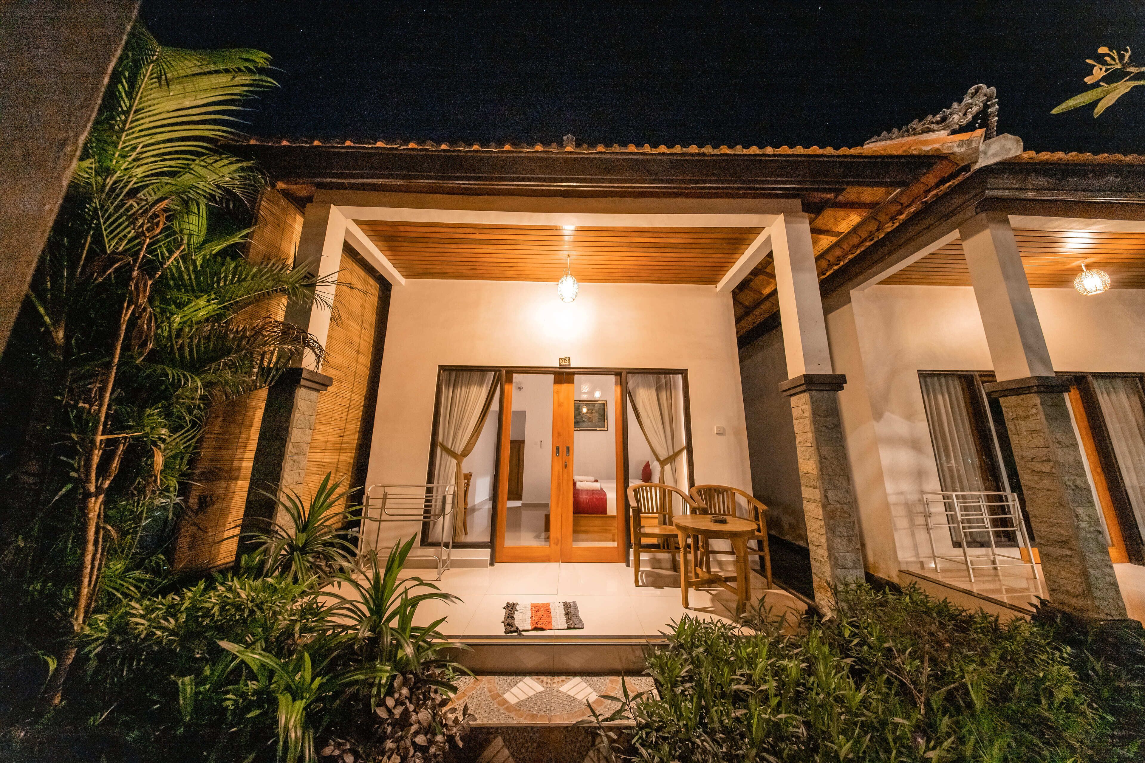A well-lit entrance with a wooden door, a small seating area with chairs, and a tiled patio.