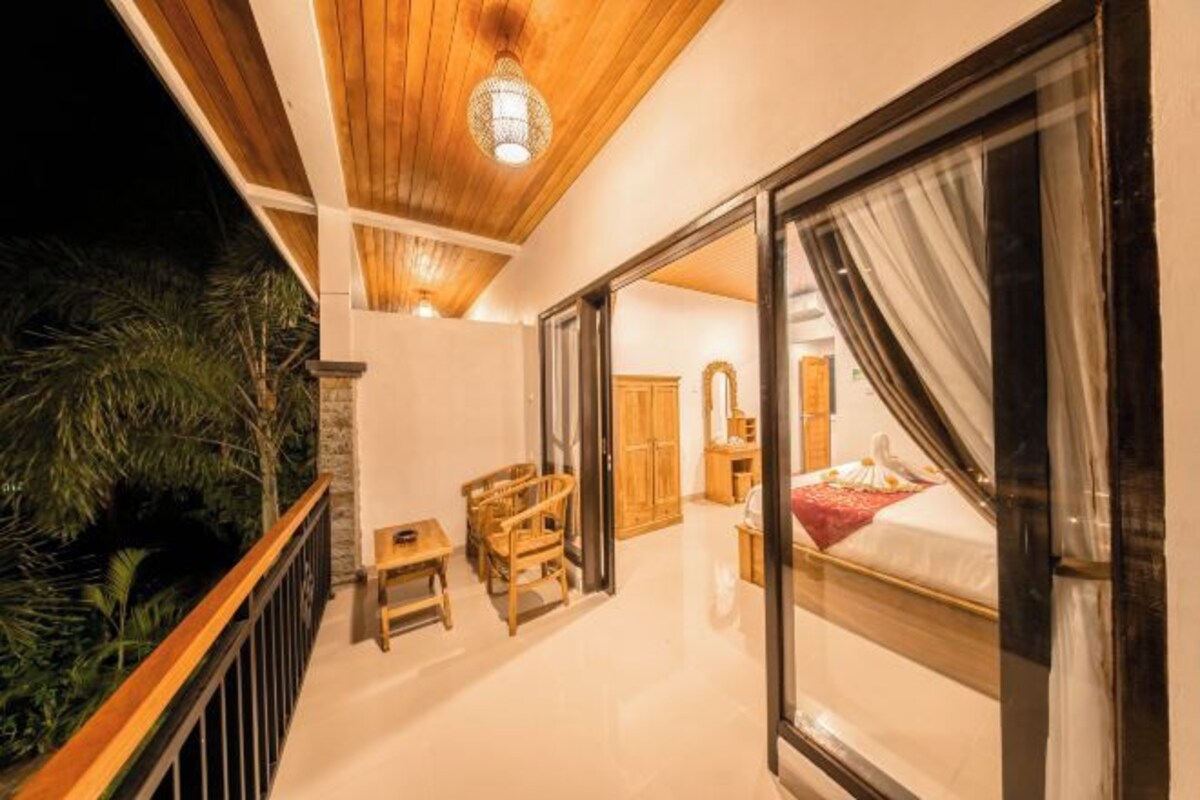 A room with a wooden ceiling, a bed with white curtains, a small table with chairs, and a view of greenery outside.