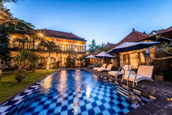 A swimming pool with checkered tiles, lounge chairs, umbrellas, and a building with a red-tiled roof.