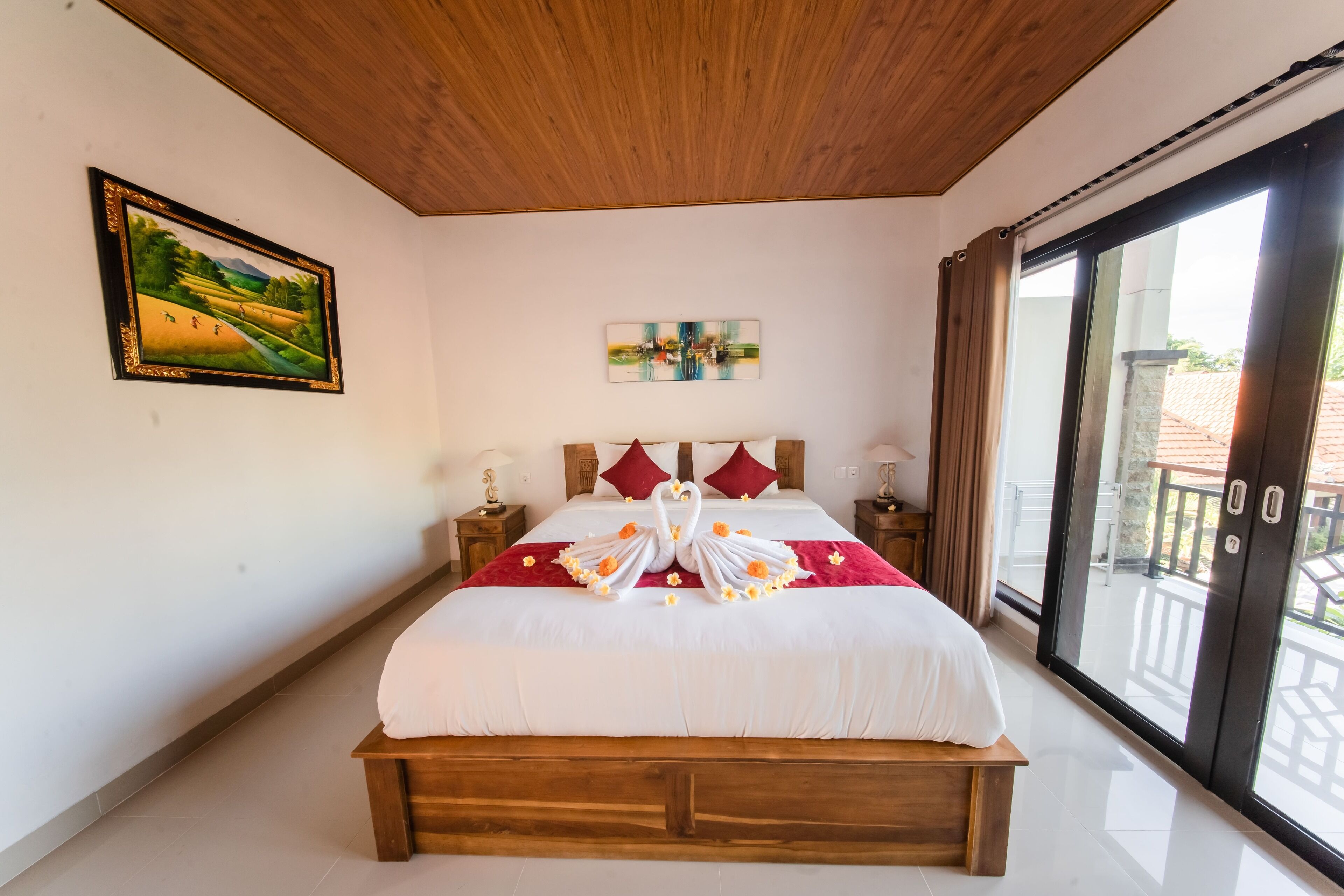 A bedroom with a wooden headboard, a bed with a red and white bedspread, a painting on the wall, and a view through a sliding glass door.