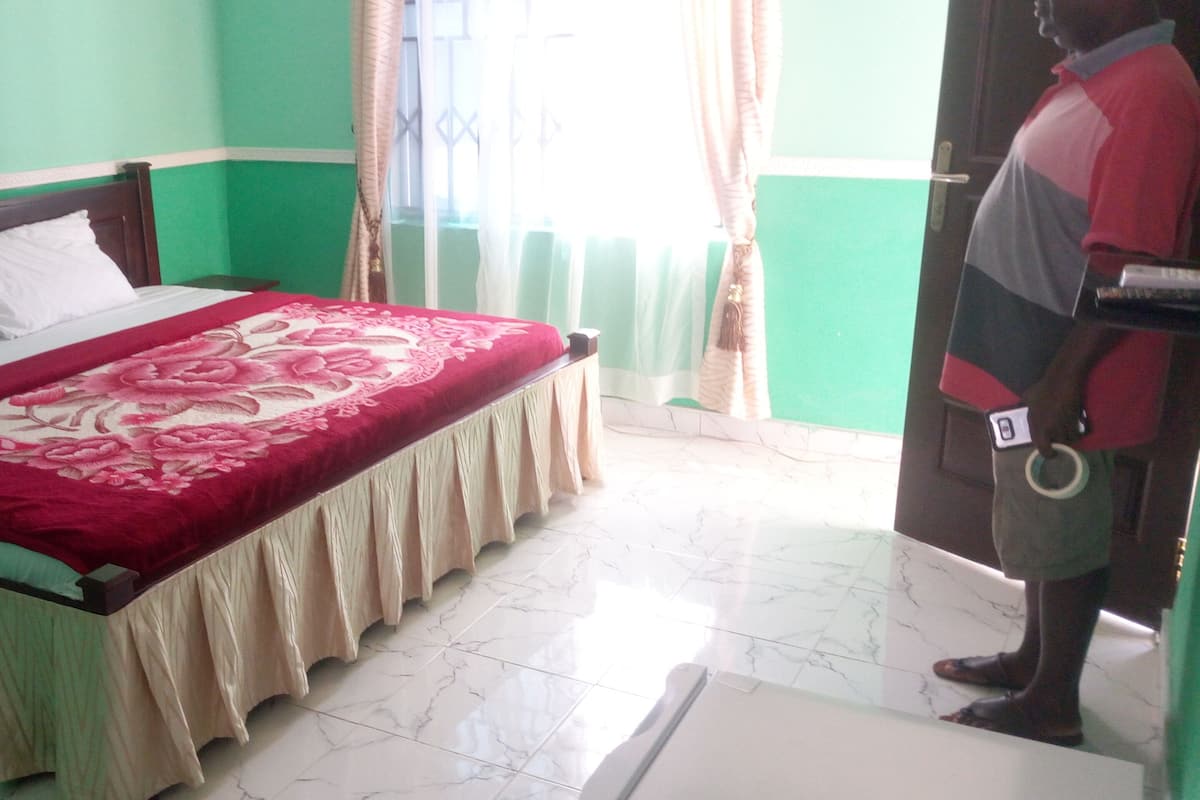 A room with a bed, green walls, a window with curtains, and a person standing by the door.
