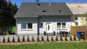 Une maison à deux étages, avec un toit recouvert de tuiles grises, une antenne parabolique et une terrasse clôturée, aménagée avec une table et des chaises.