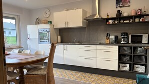 Une cuisine moderne avec des armoires blanches, une hotte de cuisinière en acier inoxydable et un four à micro-ondes. On y trouve un coin repas avec une table et des chaises, ainsi qu’une fenêtre donnant sur une maison.