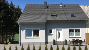 Une maison à deux étages avec un toit recouvert de tuiles grises, une porte d’entrée et deux fenêtres. On y trouve un petit balcon avec une table et des chaises.