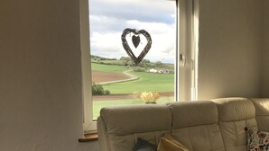 Un salon avec un canapé beige, une fenêtre décorée d’un motif en forme de cœur et une vue sur un paysage verdoyant.