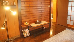 A room with wooden panel walls, a wooden floor, a small table with a basket, a magazine rack, and a floor lamp. - Yoshimura House Hotel 6 (Fujikawaguchiko)