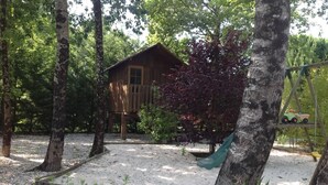 A rustic wooden cabin surrounded by trees and a gravel area with a swing set. - Le Cabassou (Begadan)