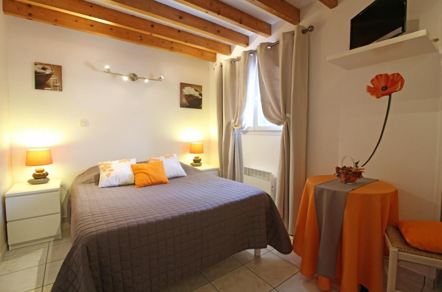 A bedroom with a bed, two bedside tables, a nightstand, a window with curtains, a small table with a chair, and a poppy decoration.