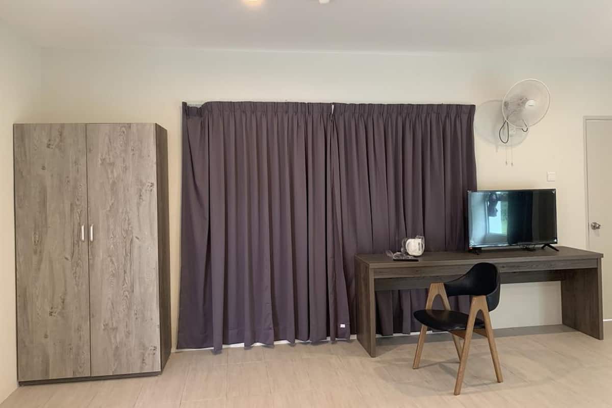 A room with a wooden wardrobe, a dark curtain, a desk with a chair, a television, and a wall-mounted fan.