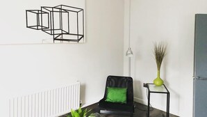 Una sala de estar moderna con un sillón de cuero negro, una mesa de centro de vidrio y un cojín verde. En la pared, hay una obra de arte abstracta enmarcada y una pequeña mesa con un florero de plantas.