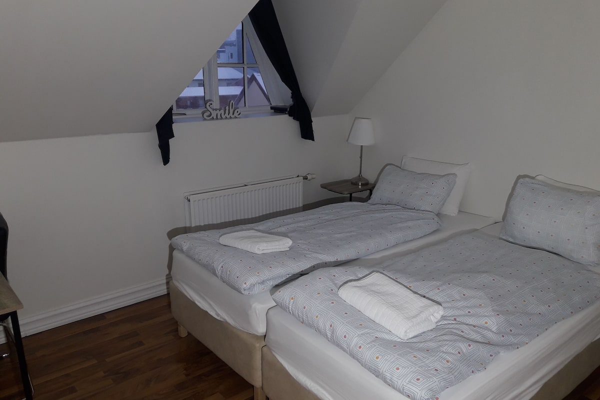 Une chambre avec deux lits, une fenêtre donnant sur des immeubles, un radiateur, une table de chevet avec une lampe et une chaise.