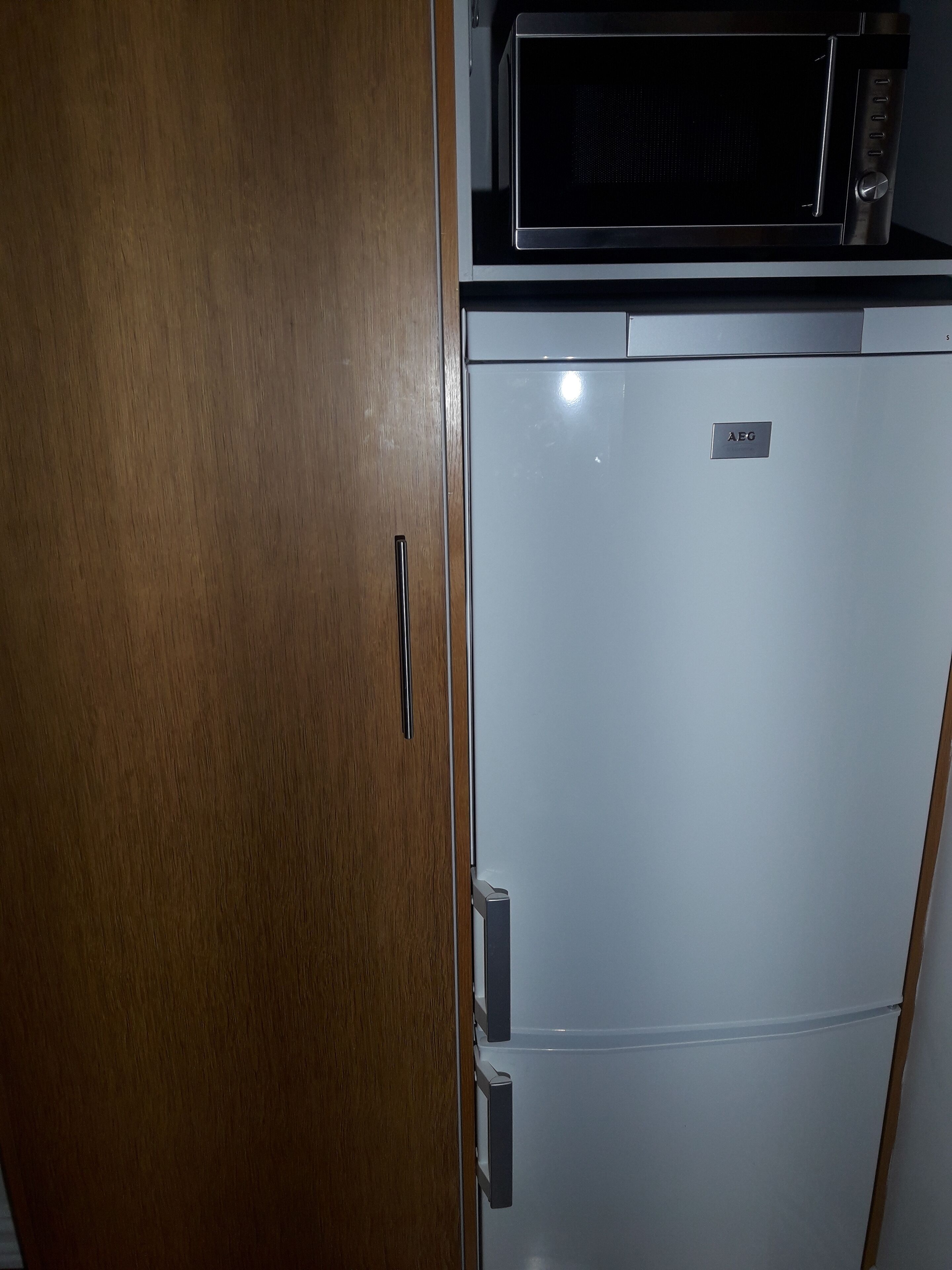 Une cuisine compacte avec un four à micro-ondes posé sur le dessus d’un réfrigérateur blanc, une porte de placard en bois et une petite étagère.