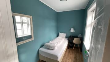 Un dormitorio pequeño y ordenado con una cama individual, un escritorio con silla, una ventana y una puerta.
