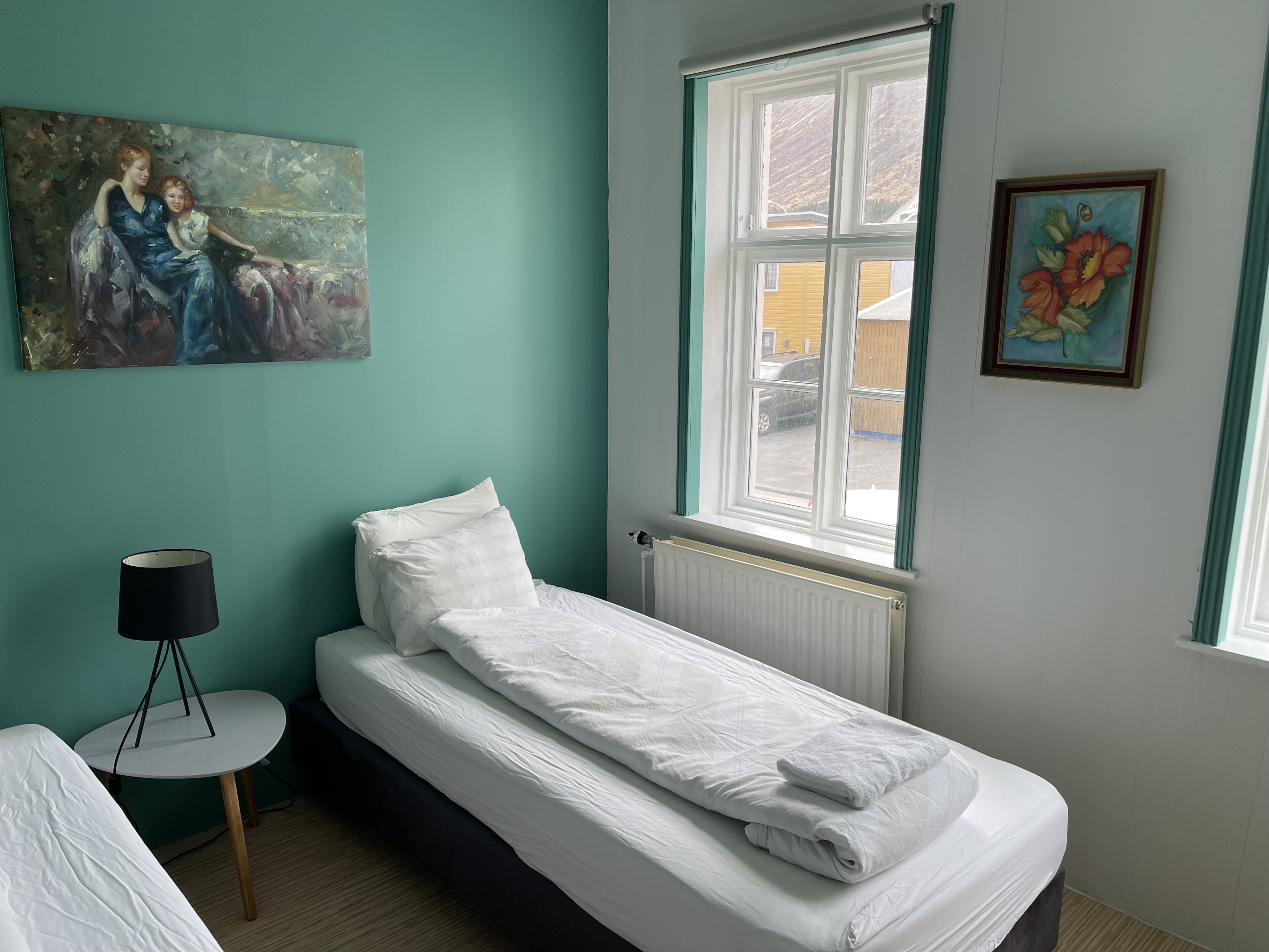 Un lit simple avec une literie blanche, une petite table de chevet avec une lampe, une fenêtre donnant sur l’extérieur et deux tableaux accrochés aux murs.