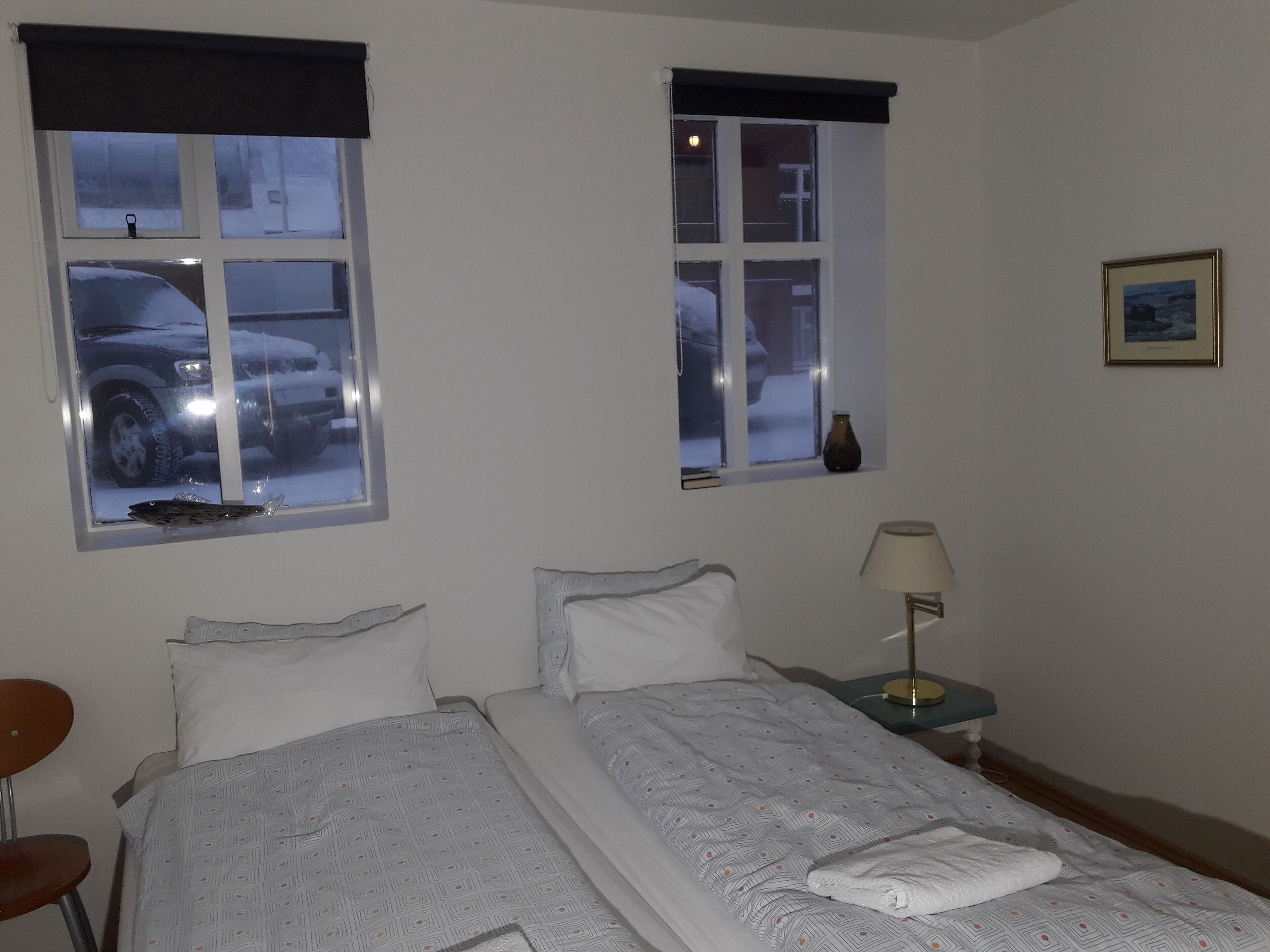 Habitación de hotel con dos camas, una silla, una mesita de noche con lámpara y dos ventanas con vistas a una calle nevada.