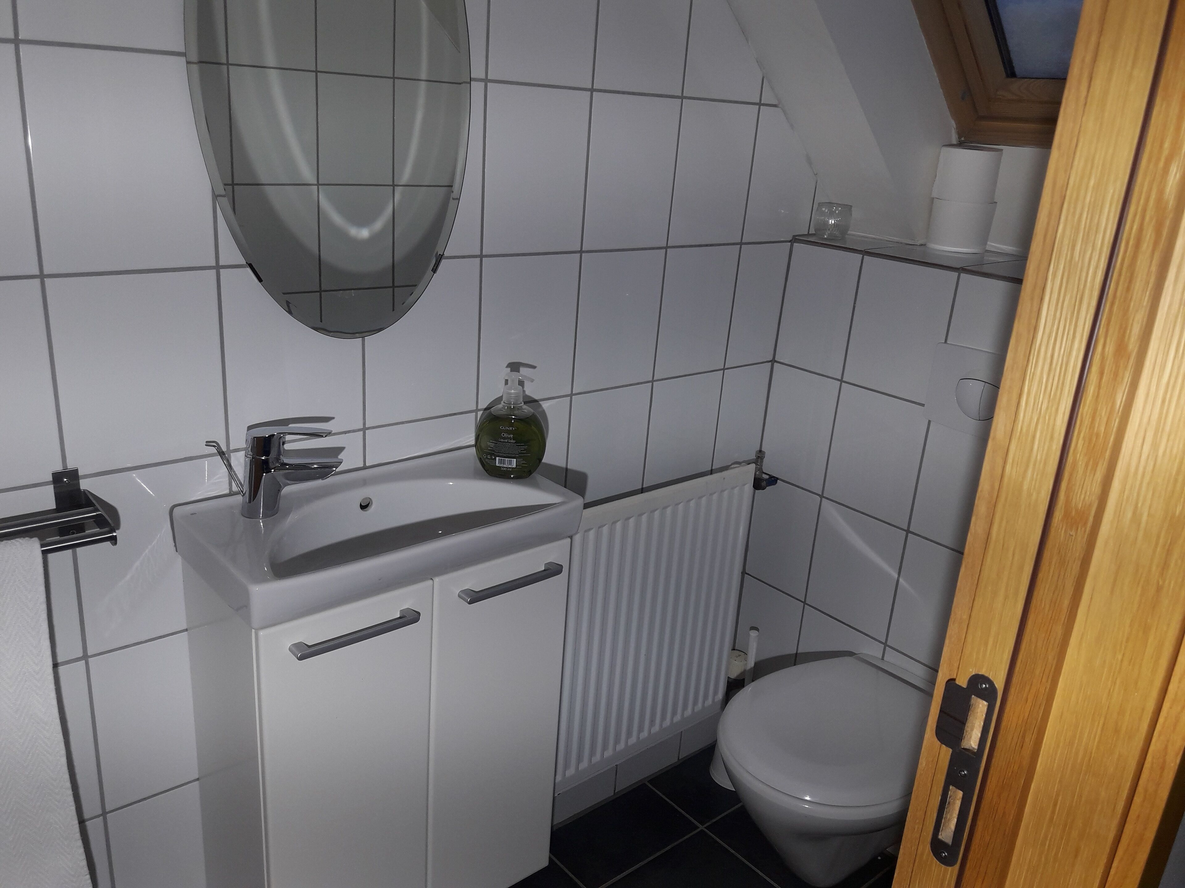 A small, clean bathroom with a white sink, mirror, and toilet. There is a radiator under the sink and a shelf above the toilet.