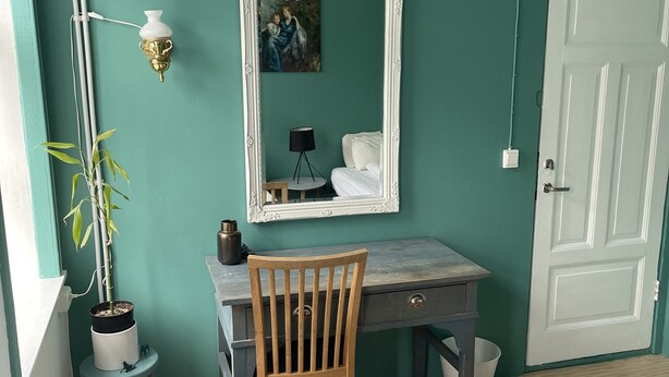 Ein Zimmer mit türkisfarbenen Wänden, einer weißen Tür, einem Schreibtisch mit Stuhl, einem Bett mit weißer Decke, einer Zimmerpflanze und einer Wandlampe.