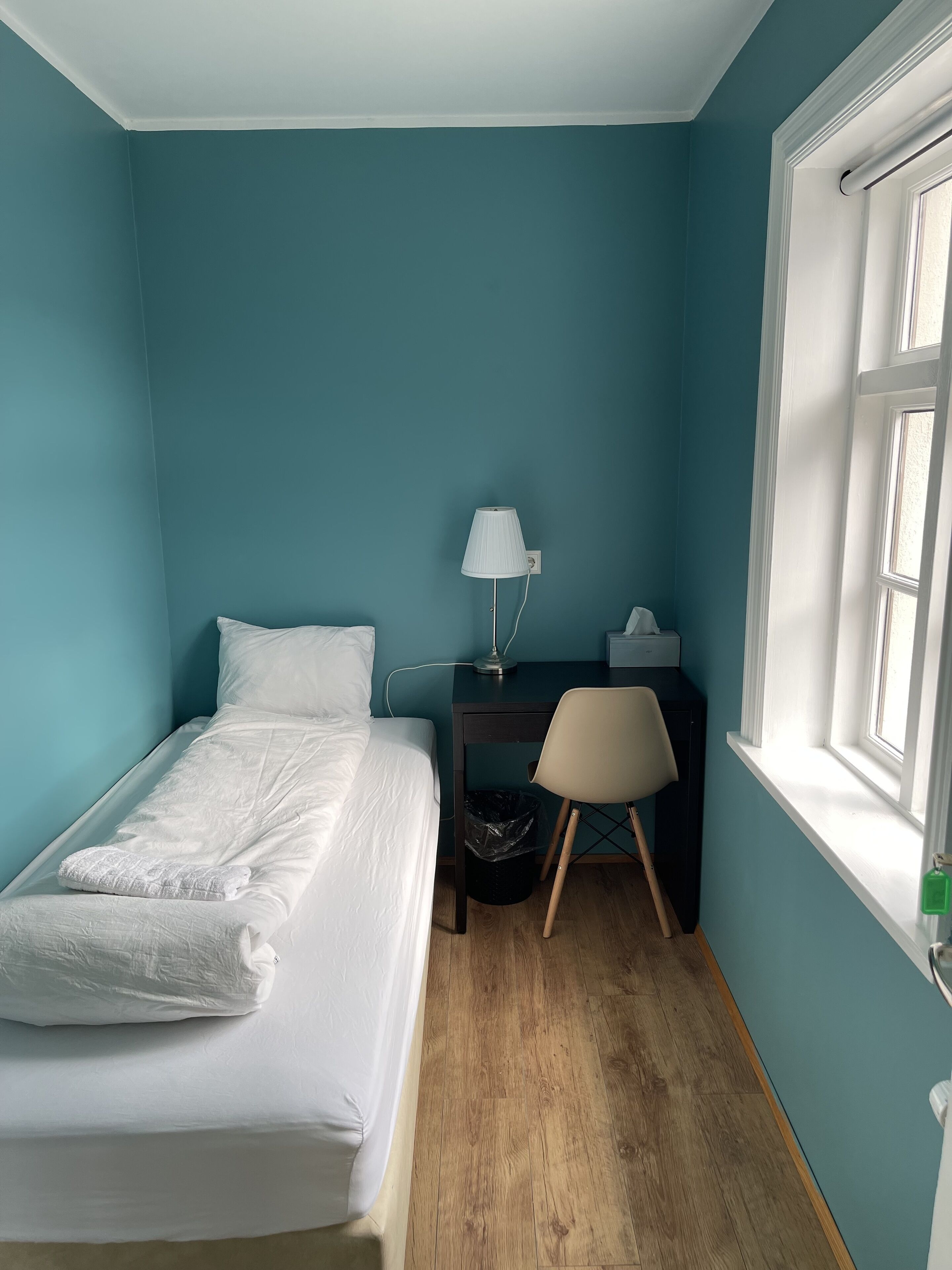A bedroom with teal walls, a bed with white linens, a desk with a lamp, and a chair.