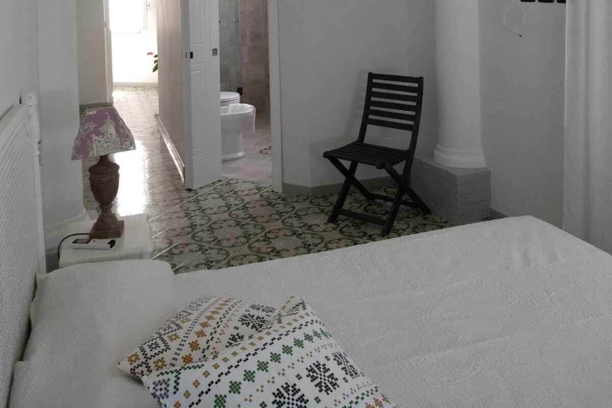 A bedroom with a bed, a chair, a chandelier, and a bathroom visible through an archway.
