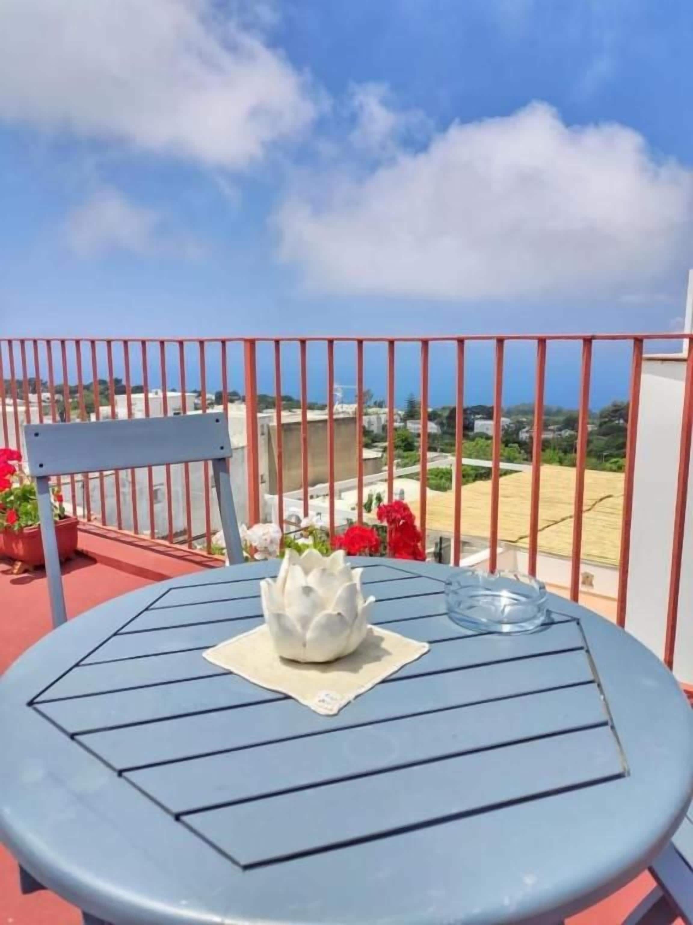 A round table with a white decorative item on a blue tablecloth, set against a red railing with a view of a beach and buildings in the background.