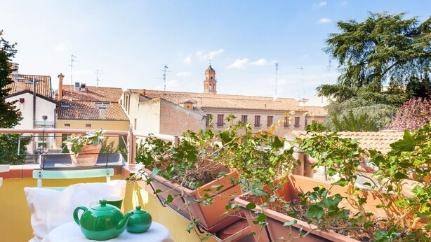 Une terrasse sur le toit agrémentée de jardinières en terre cuite, une table avec une théière verte, et une vue sur des immeubles avec une tour en arrière-plan.