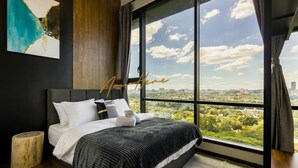 Quarto moderno com uma cama grande, vista da cidade e uma obra de arte abstrata na parede.