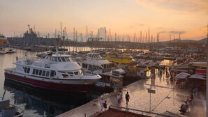 A marina with boats docked, a pedestrian walkway, and a sunset in the background.