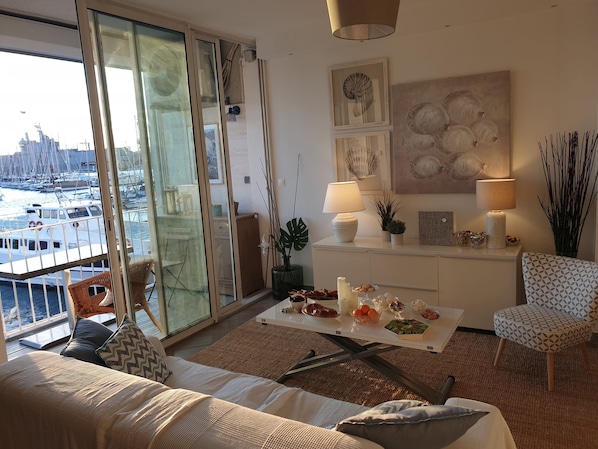 Un salon moderne avec vue sur un port de plaisance, une porte vitrée donnant sur l’extérieur, un canapé blanc, une table basse avec des collations et un fauteuil à motifs.