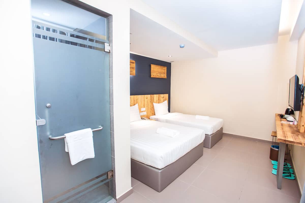 A hotel room with two beds, a wooden headboard, a small desk, and a glass shower enclosure.
