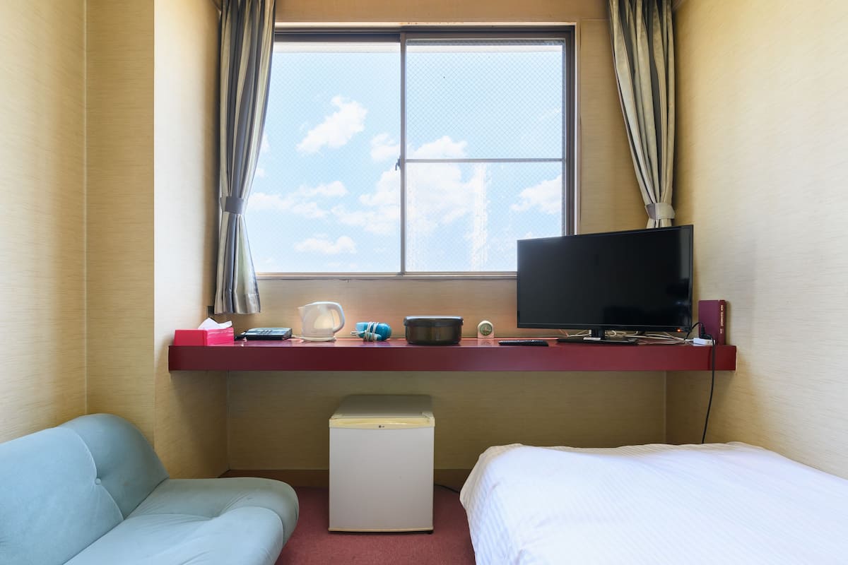 A hotel room with a bed, a desk with a TV, a small fridge, a window with curtains, and a red shelf with various items.