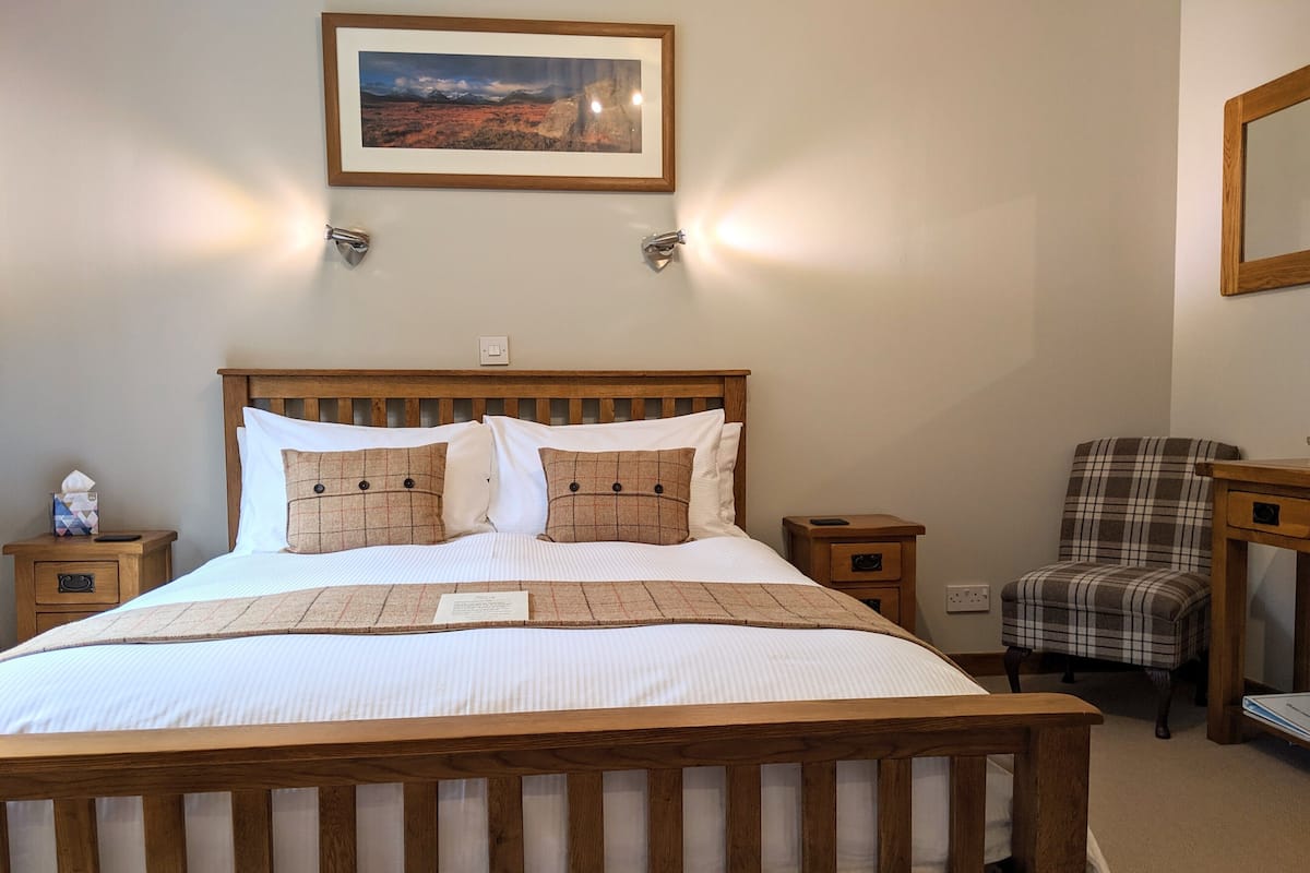 A neatly made bed with a wooden headboard, a plaid armchair, and a framed picture on the wall.