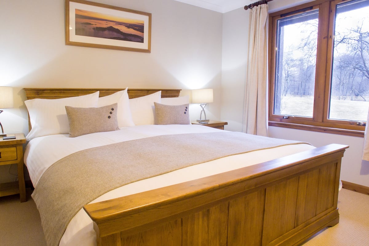 A neatly made bed with a wooden headboard, two bedside tables with lamps, a framed picture on the wall, and a window with curtains.