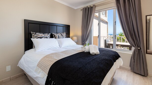 Een netjes opgemaakt bed met een zwart hoofdeinde, witte en gemønsterde kussens en een beige en zwarte deken. Er is een raam met gordijnen en uitzicht op de omgeving.