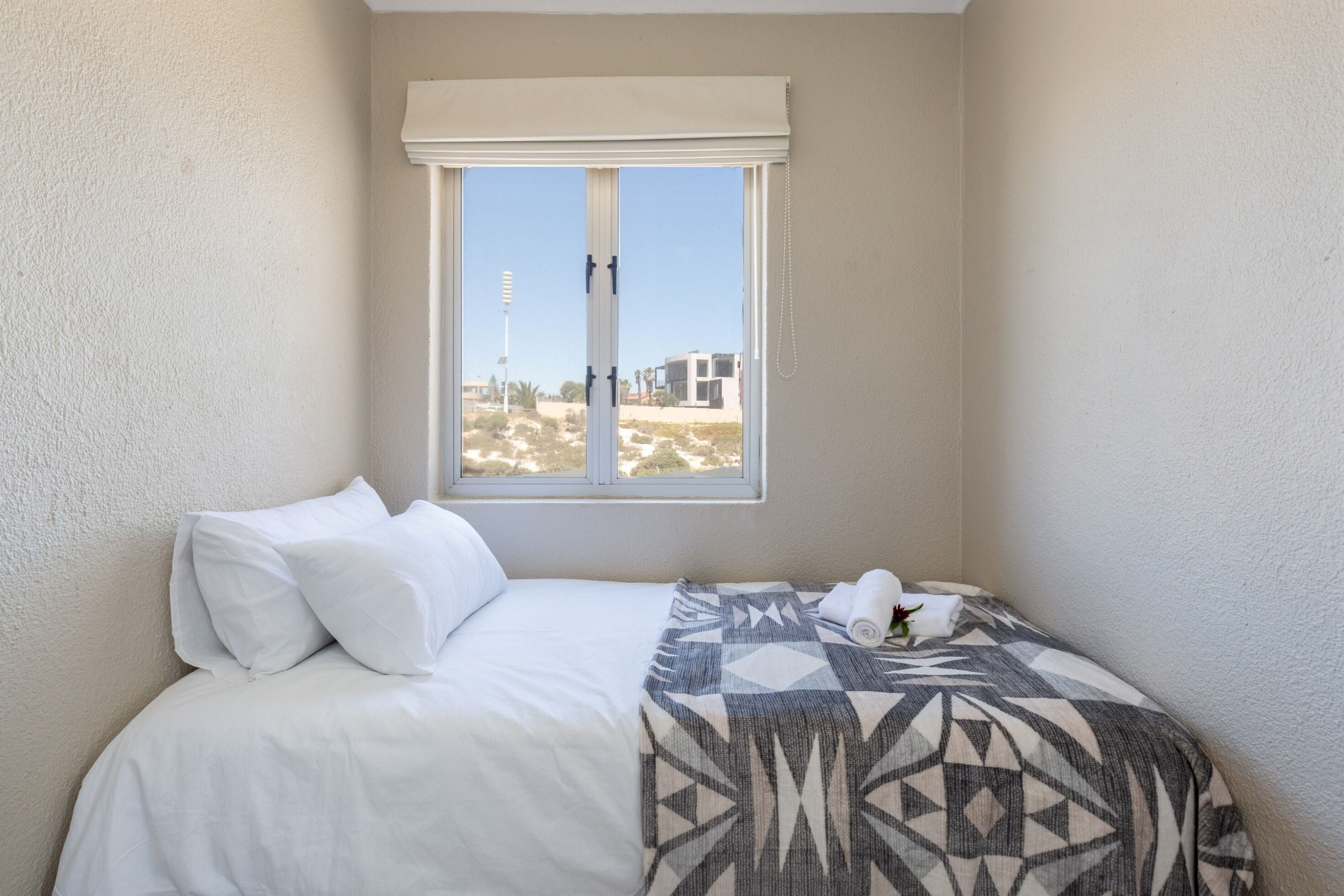 A neatly made bed with white bedding and a patterned throw blanket, a window with a view of buildings and a clear sky, and a white wall.