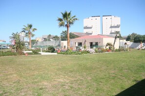 Il y a une zone herbeuse avec un bâtiment doté d’un toit rouge et de plusieurs étages. Des palmiers et des plantes en pot sont disposés autour du bâtiment.