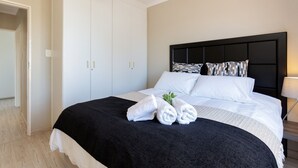 Een netjes opgemaakt bed met witte lakens, een zwarte plaid en opgerolde handdoeken. Daarboven hangt een plafondventilator en rechts van het bed staat een nachtkastje met een lamp.