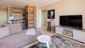 Een moderne woonkamer met een bank, een glazen salontafel, een flatscreen televisie en een keukenhoek met kasten en apparatuur.