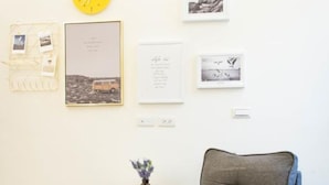 A room with a grey armchair, a small white side table, a yellow clock, and framed pictures on the wall.