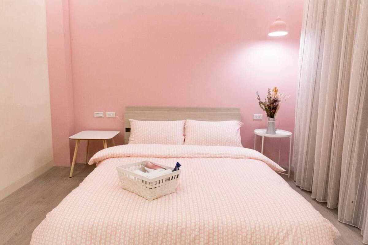 A bedroom with a pink wall, a bed with a white and pink patterned bedspread, a small bedside table, a nightstand with a vase of flowers, and a window with curtains.