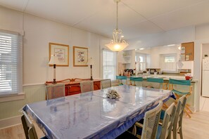 A dining area with a covered table, chairs, a chandelier, framed artwork, and a kitchen in the background.