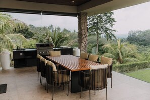 An outdoor dining area with a wooden table and chairs, a grill, and a view of lush greenery. - White Monkey Villa - Private Pool & Jacuzzi (Langkawi)