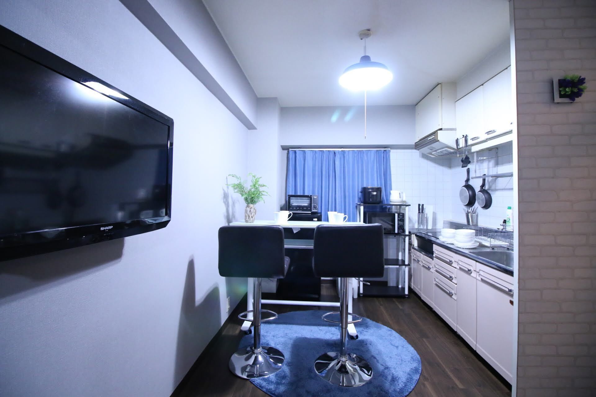Uma cozinha compacta com televisão de tela plana, uma área de refeições com duas cadeiras, micro-ondas, chaleira e cortina azul.