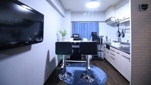 A compact kitchen with a flat-screen TV, a dining area with two chairs, a microwave, a kettle, and a blue curtain.