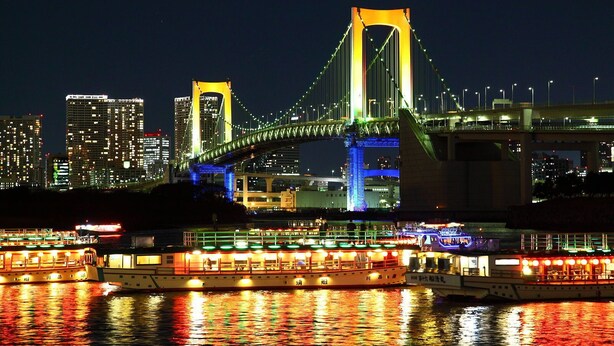 A brightly lit bridge with two towers and a river with boats.