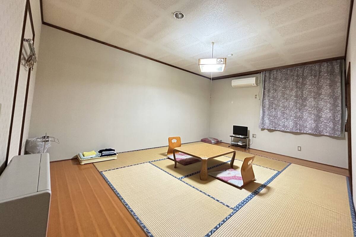 Een kamer met tatami vloerbedekking, een salontje, een stoel, een televisie en een raam met gordijn.