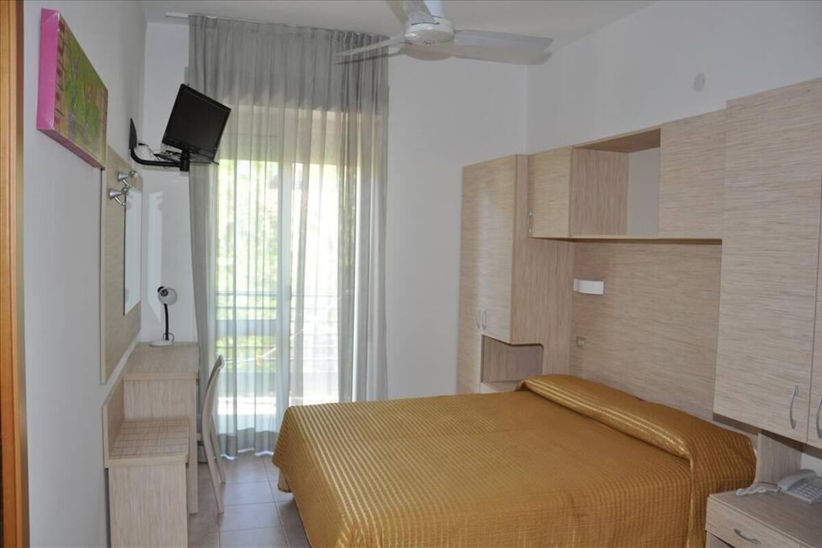 A bedroom with a bed, a TV mounted on the wall, a ceiling fan, and a window with curtains.