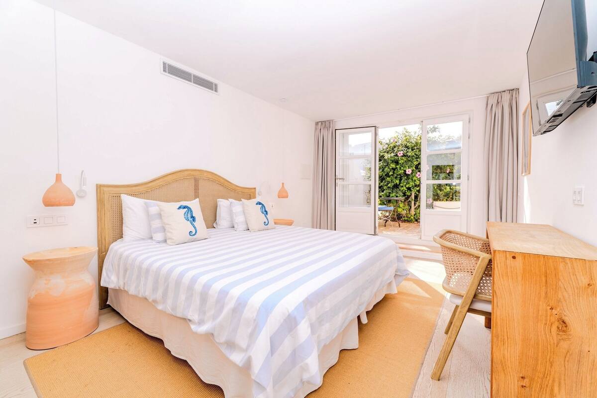 Quarto com cama grande, cabeceira de madeira, cadeira de vime e pequena mesa.