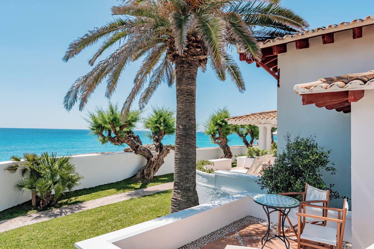 Um pátio com vista para o mar, uma palmeira e um edifício branco com telhado de azulejos.