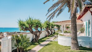 A coastal property with a pathway leading to a white building with green shutters, surrounded by palm trees and a view of the sea. - The Sea Club (Capdepera)