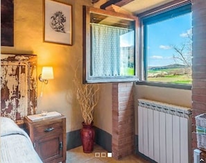 A room with a window overlooking a landscape, a radiator, a wooden nightstand with a lamp, and a vase with branches.
